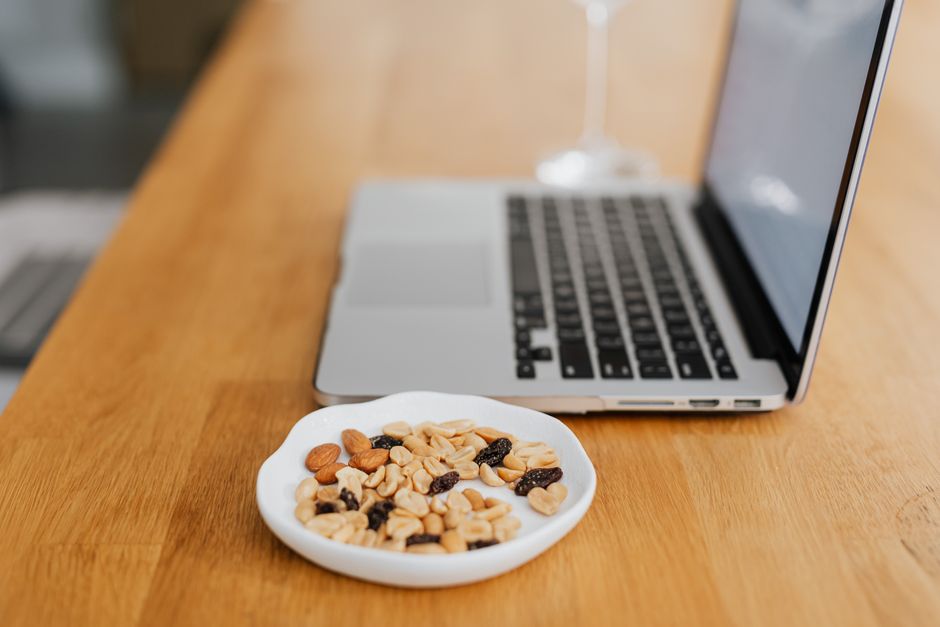 Imagem de um ambiente de trabalho com laptop aberto e uma tigela de mix de castanhas e frutas secas sobre uma mesa de madeira, transmitindo um momento de produtividade aliado ao cuidado com a alimentação.
