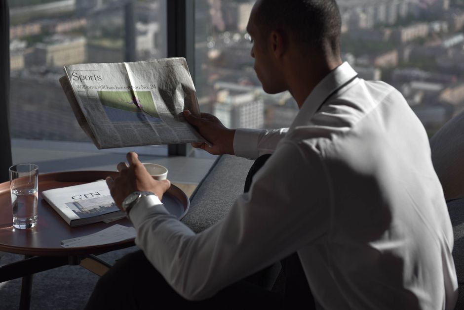 Homem lendo jornal em ambiente corporativo, com vista urbana ao fundo, transmitindo uma atmosfera de negócios e atualização profissional.