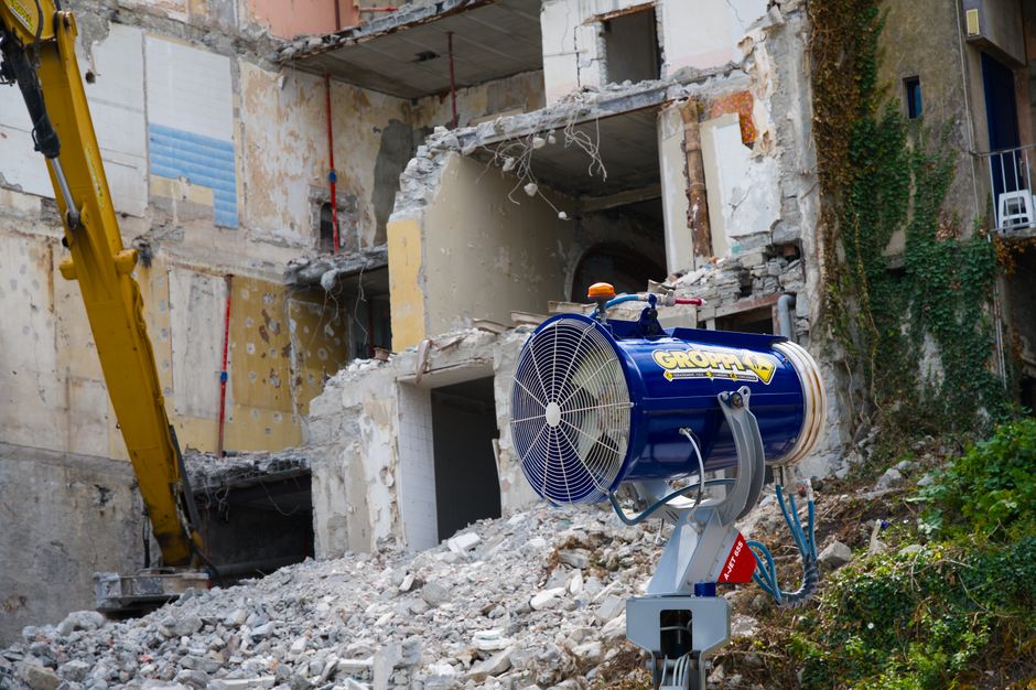 Imagem de uma construção ou demolição em andamento, com uma máquina de circulação de ar em primeiro plano e uma estrutura de prédio parcialmente destruída ao fundo.