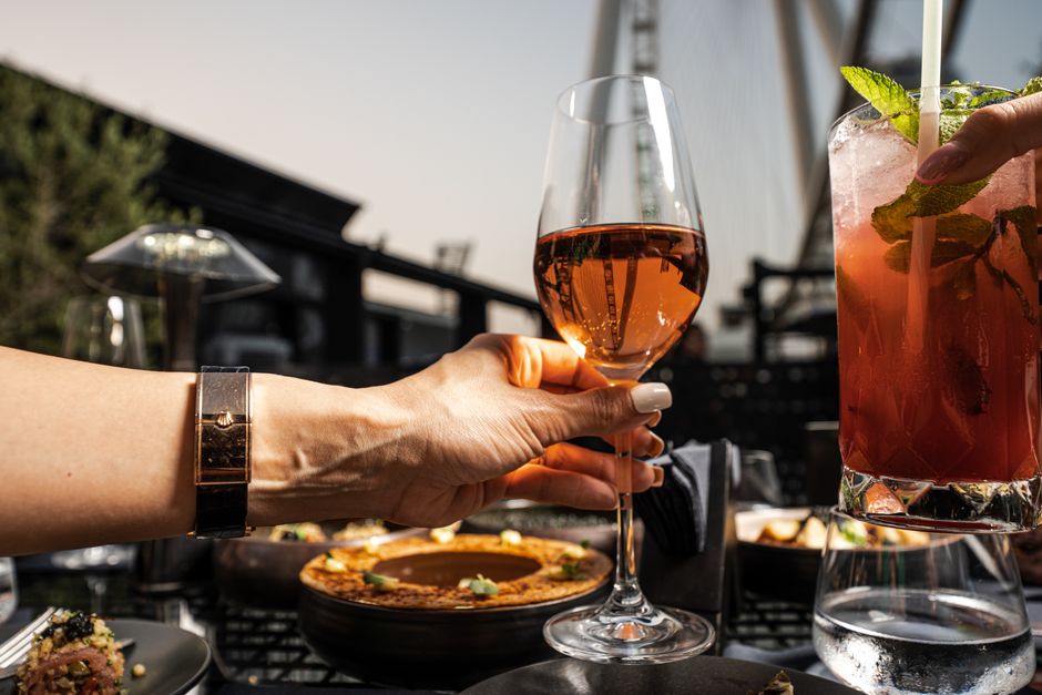 Pessoas brindando com drinks e pizza em um ambiente ao ar livre ao entardecer, criando uma atmosfera de confraternização e lazer.