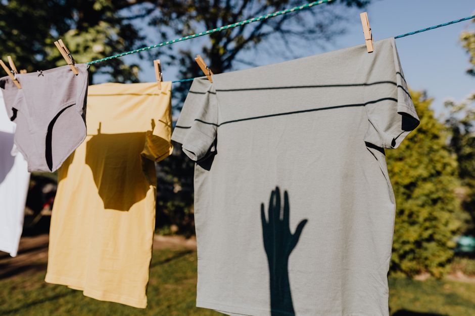 Roupas penduradas em uma linha de varal ao ar livre, com sombra de uma mão projetada na camiseta cinza, transmitindo uma atmosfera de cotidiano e criatividade ao ambiente externo.