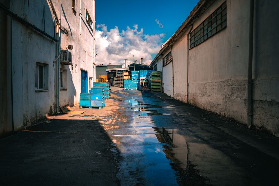 Imagem de um pátio industrial com céu claro e várias pilhas de caixas e pallets ao fundo.
