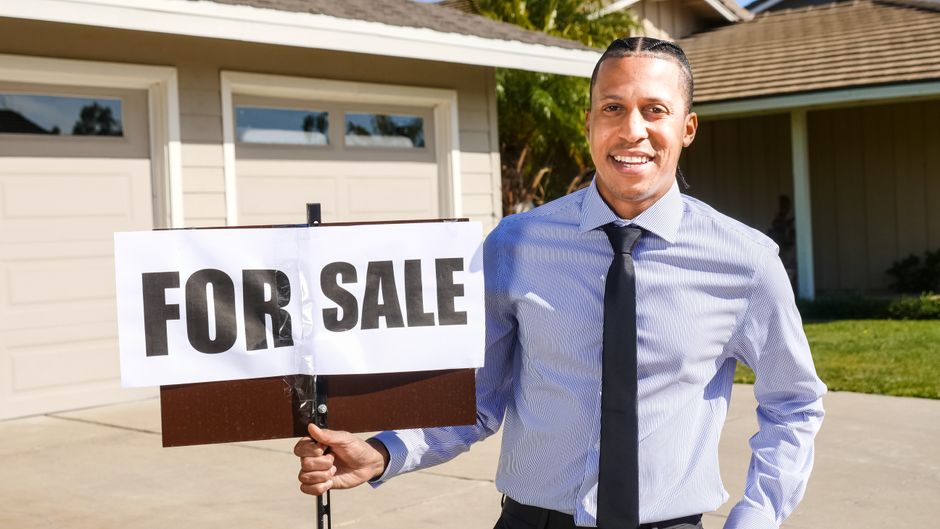 Homem sorridente ao lado de um cartaz de 'Para Venda', representando o tema de venda de imóveis ou negócios residenciais.