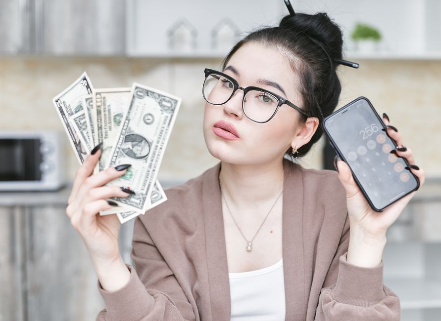 Jovem mulher com expressão irônica segurando dinheiro e celular, refletindo sobre finanças pessoais ou despesas.