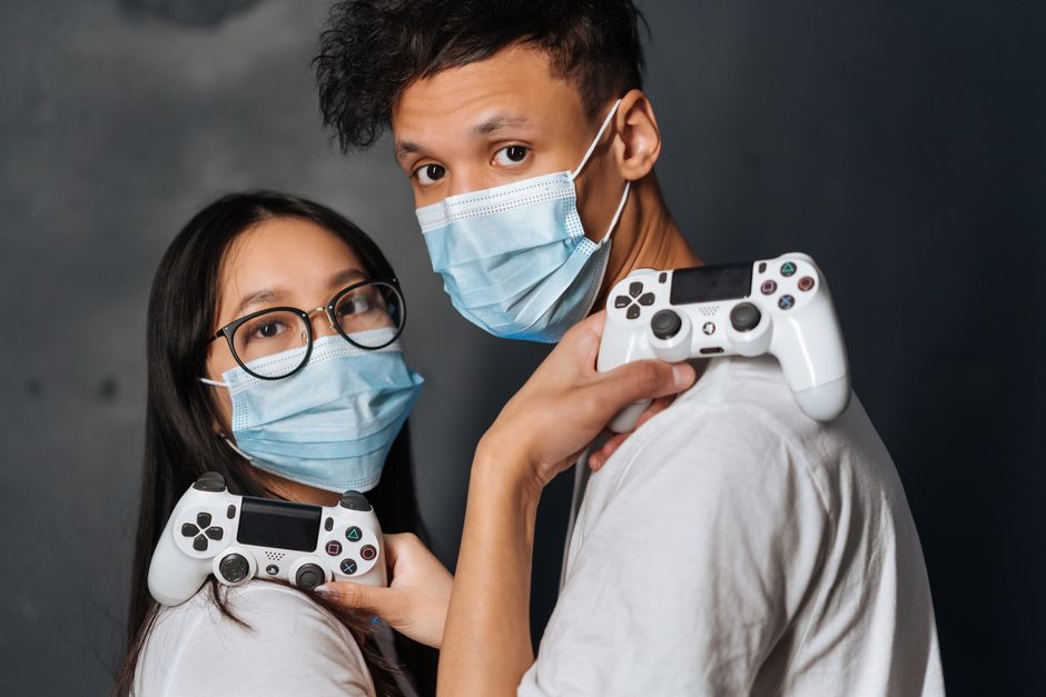 Imagem de um jovem casal usando máscara facial e segurando controles de videogame, representando diversão segura em tempos de pandemia.