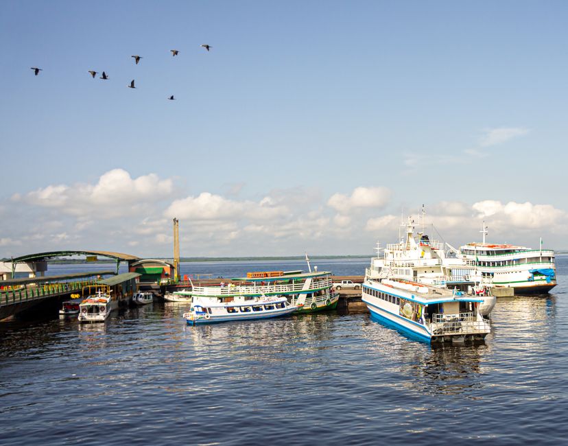 Imagem de um porto com vários barcos e navios atracados sob um céu ensolarado, evocando a temática de viagens, transporte marítimo e turismo.
