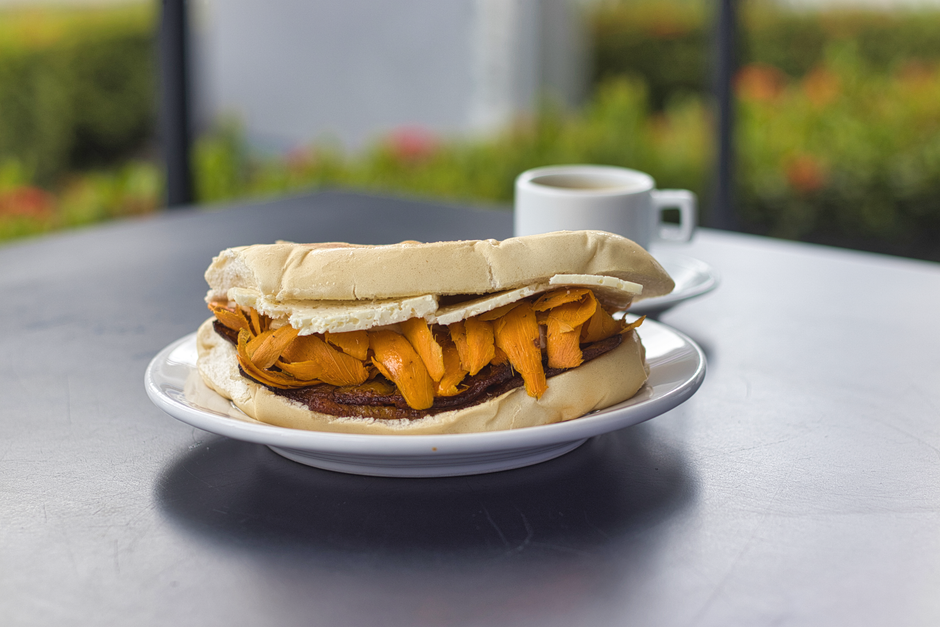 Sanduíche de pão francês com recheio de carne e abóbora, acompanhado de uma xícara de café em ambiente externo.