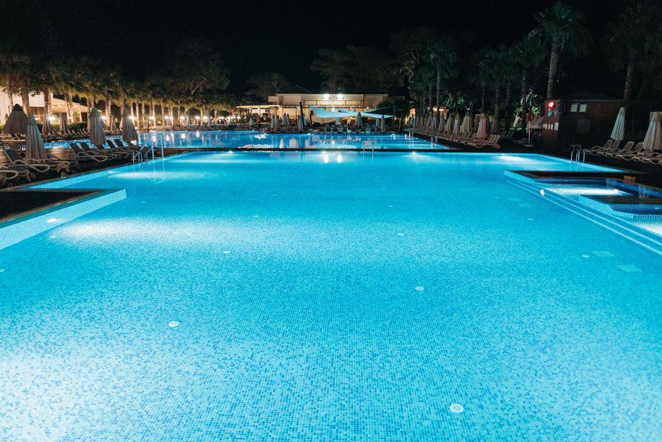 Piscina iluminada à noite em um ambiente de resort ou hotel de luxo, com espreguiçadeiras e área de lazer ao redor.