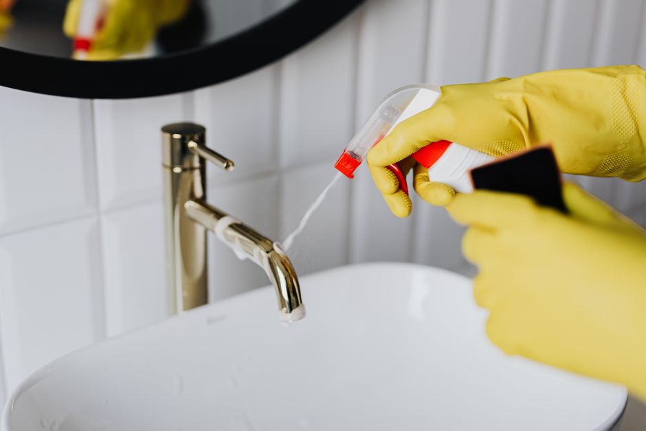 Pessoa usando luvas amarelas e spray desinfetante para higienizar uma pia de banheiro, reforçando a importância da limpeza e sanitização em ambientes públicos ou profissionais.