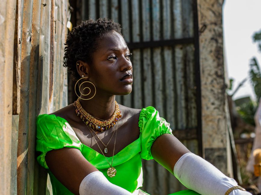 Mulher africana com expressão pensativa sentada ao lado de uma parede de madeira, vestindo roupa vibrante e acessórios coloridos, refletindo sobre temas culturais ou sociais.