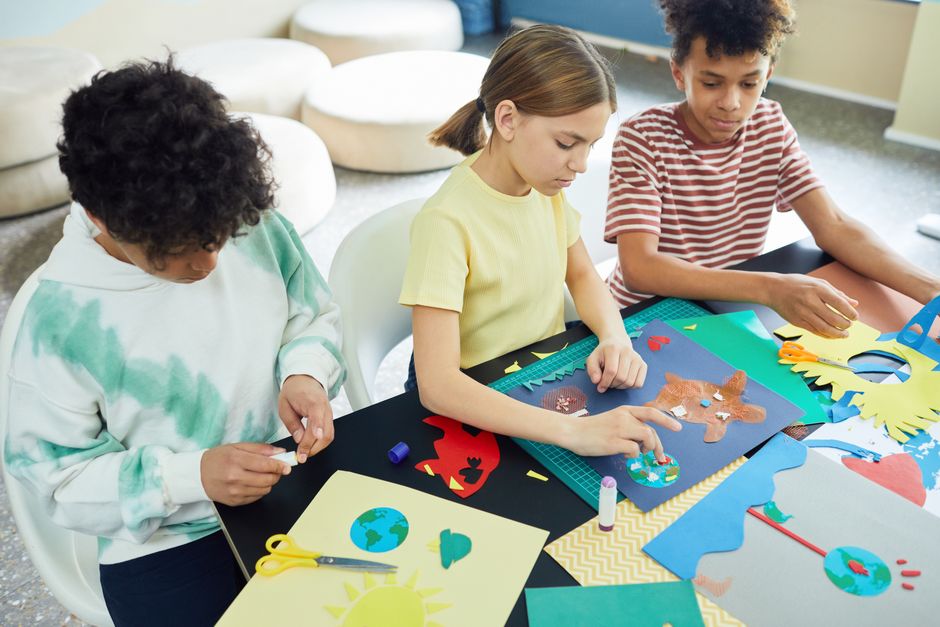 Três crianças participando de uma atividade criativa de artesanato com recortes coloridos e papel.