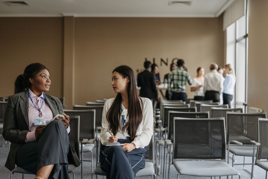 Duas mulheres conversando em uma sala de conferências com várias pessoas ao fundo, sugerindo networking ou discussão profissional.