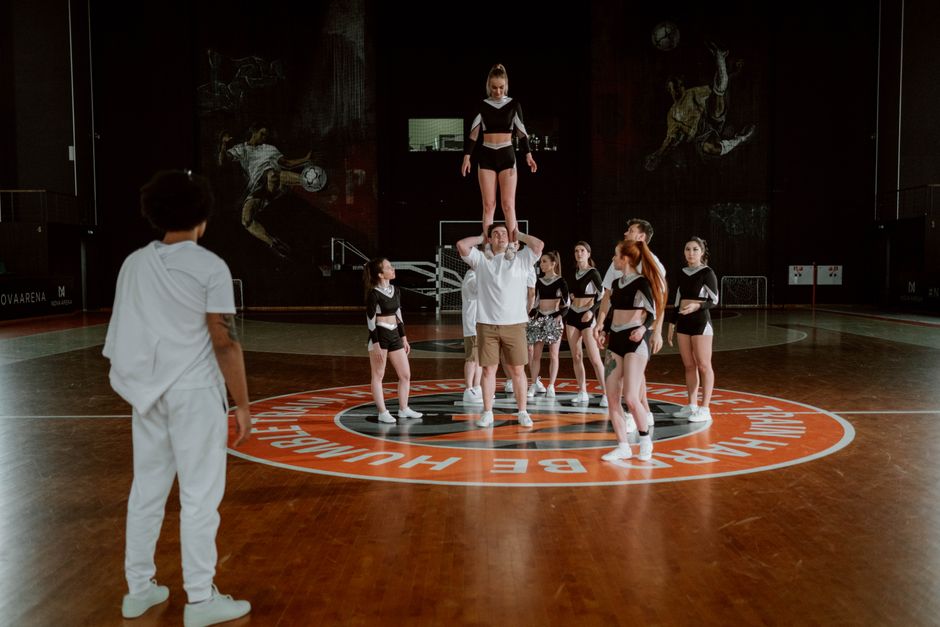 Grupo de jovens praticando uma atividade de ginástica artística no indoor, com uma menina equilibrada em um supervisor e uma observadora ao lado, em um ambiente esportivo com piso de ginásio.