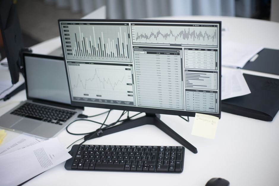 Imagem de uma mesa de trabalho com computador exibindo gráficos financeiros e planilhas, sugerindo uma análise de dados ou gestão financeira.