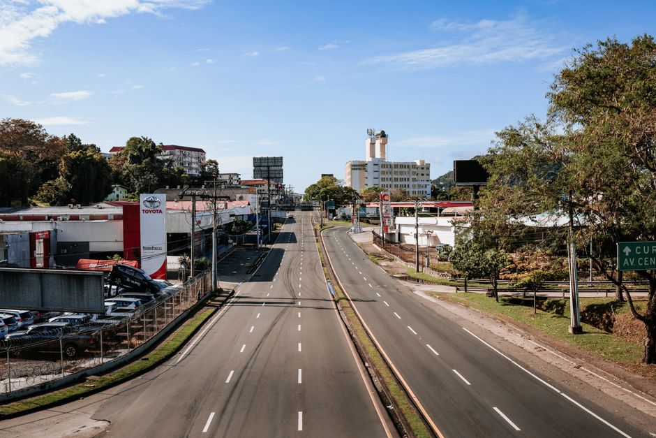 Imagem de uma rua urbana vazia, com edifícios comerciais e árvores ao redor, sob um céu claro, destacando a quietude em um dia ensolarado.