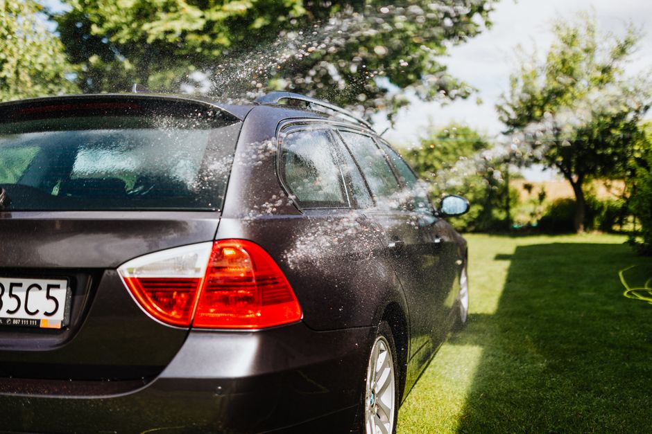 Imagem de um carro preto sendo lavado ao ar livre em um jardim com árvores e grama, destacando a limpeza de veículos.