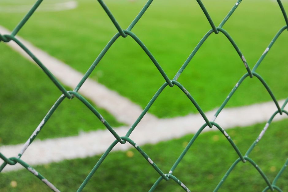 Imagem de uma cerca de arame, com um campo de futebol ao fundo, destacando a separação entre zonas de uso e restrição.