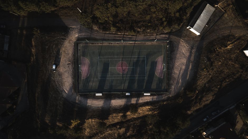 Imagem aérea de uma quadra de esportes coberta, rodeada por árvores e áreas de terra, capturada ao entardecer ou amanhecer.