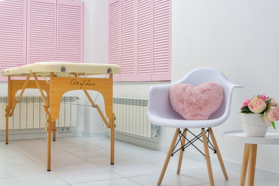 Ambiente de clínica ou spa com atmosfera acolhedora, apresentando uma cadeira com almofada de coração, uma mesa de apoio com flores e um macrobiótico na parede, transmitindo conforto e cuidado à saúde e bem-estar.