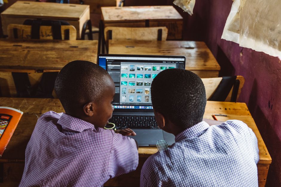 Dois estudantes negros trabalhando juntos em um computador portátil em um ambiente de sala de aula, focados em pesquisa ou projeto escolar.