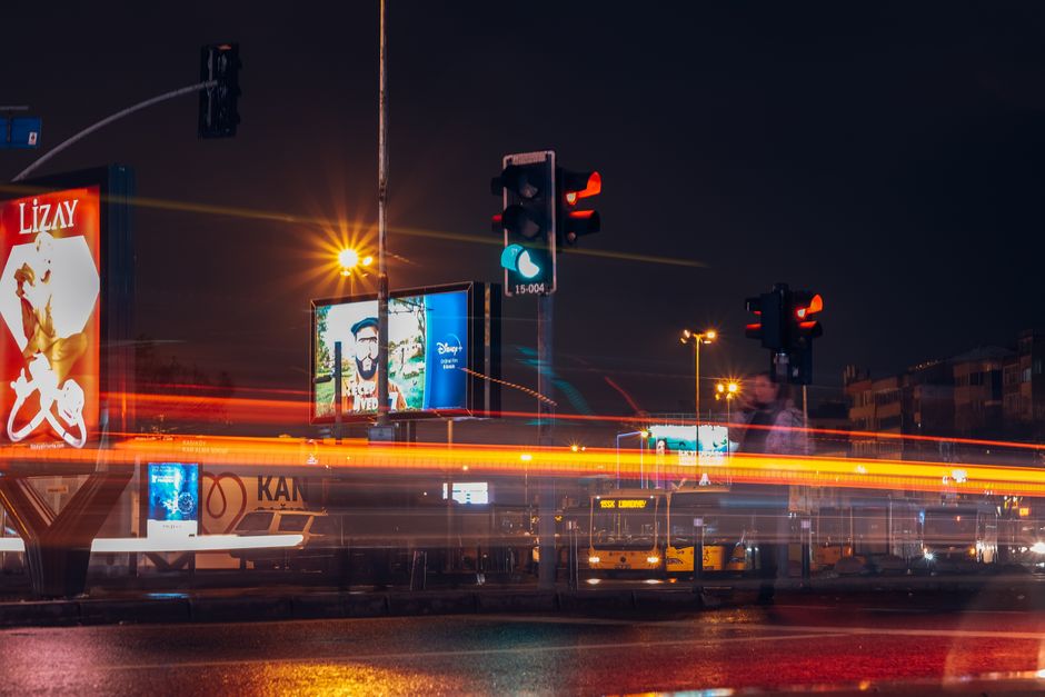 Foto de uma esquina movimentada à noite com semáforos, outdoors iluminados e luzes de veículos em movimento, destacando o cenário urbano noturno.