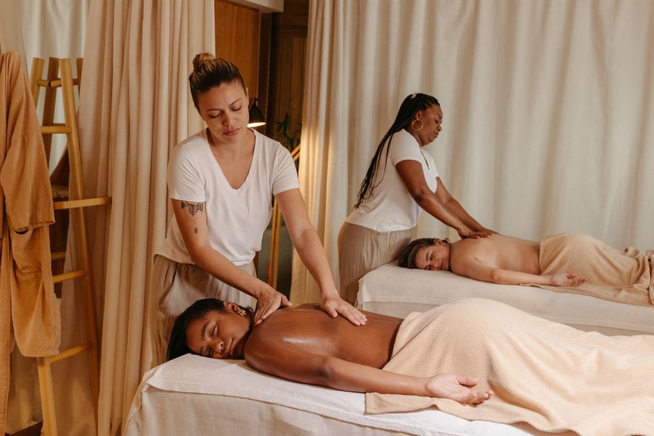 Duas terapeutas realizando sessões de massagem relaxante em um ambiente de bem-estar com cortinas clarinhas e atmosfera acolhedora.