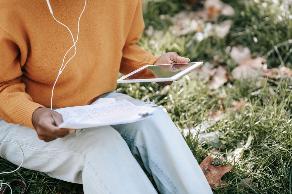 Pessoa realizando estudos ou trabalho ao ar livre utilizando dispositivo digital e materiais impressos, em um ambiente natural e tranquilo.