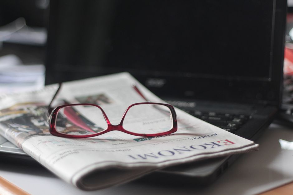 Óculos de leitura sobre um jornal na frente de um laptop, indicando foco em notícias e análise de informações financeiras ou de negócios.