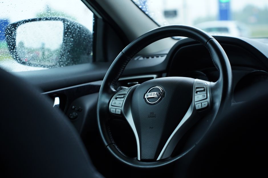 Imagem de um carro no trânsito durante um dia chuvoso, capturada do interior do veículo com foco no volante e na janela molhada.