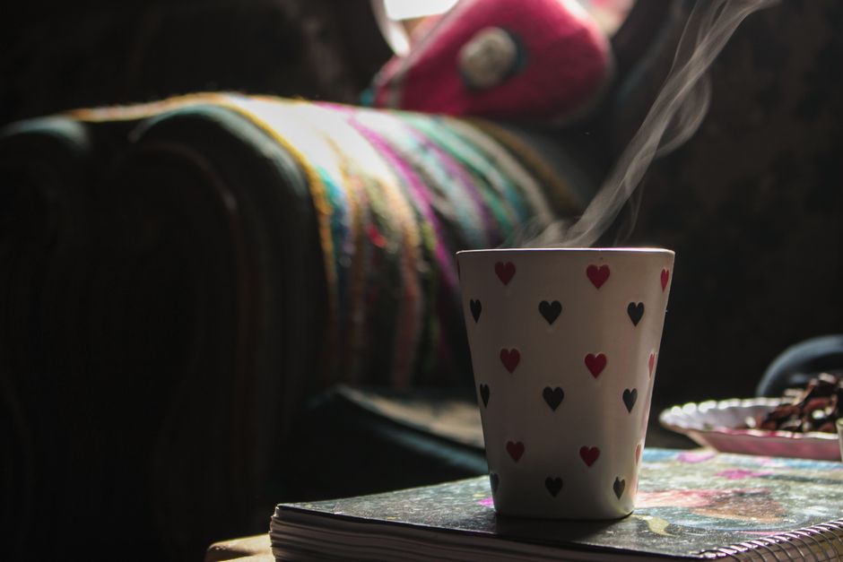 Imagem de uma xícara de café com fumaça, sobre uma mesa ao lado de um caderno ou revista, com uma manta colorida ao fundo, transmitindo aconchego e momento de pausa.