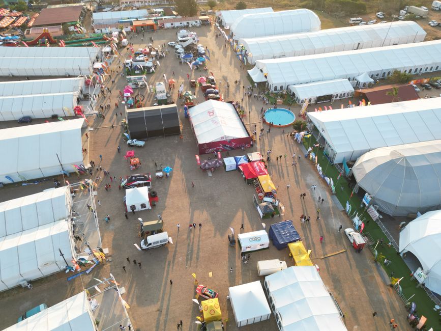 Imagem aérea de um evento ao ar livre com tendas, várias pessoas, veículos e áreas de convivência, sugerindo um festival ou feira de grande porte.