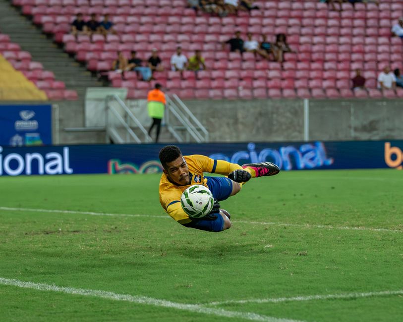 Goleiro em ação esticando-se para defender a bola durante uma partida de futebol em estádio com público ao fundo.