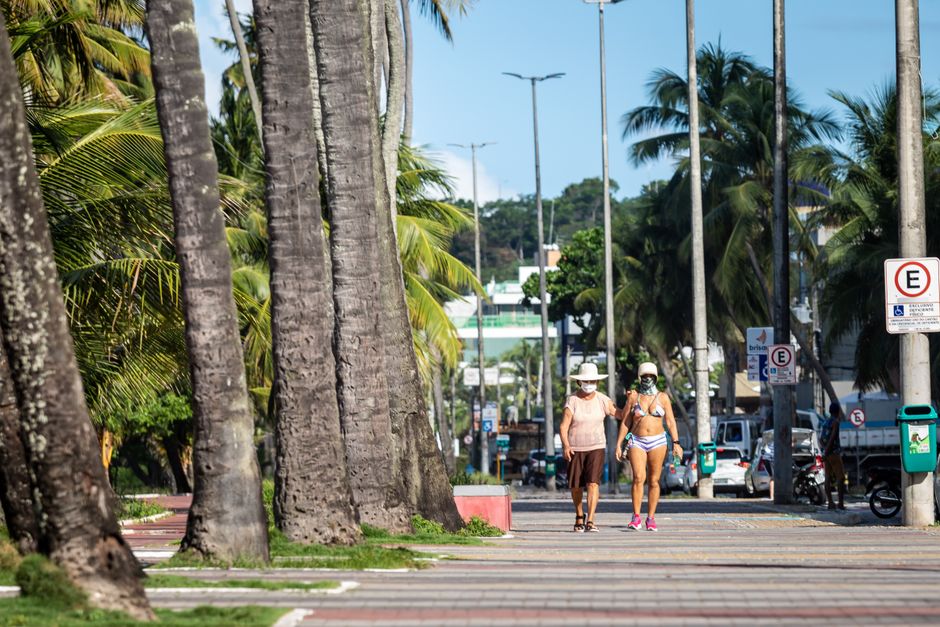 Duas mulheres caminhando na orla de uma cidade tropical, usando roupas de banho e chapéus, sob a sombra de coqueiros e ao lado de uma avenida movimentada