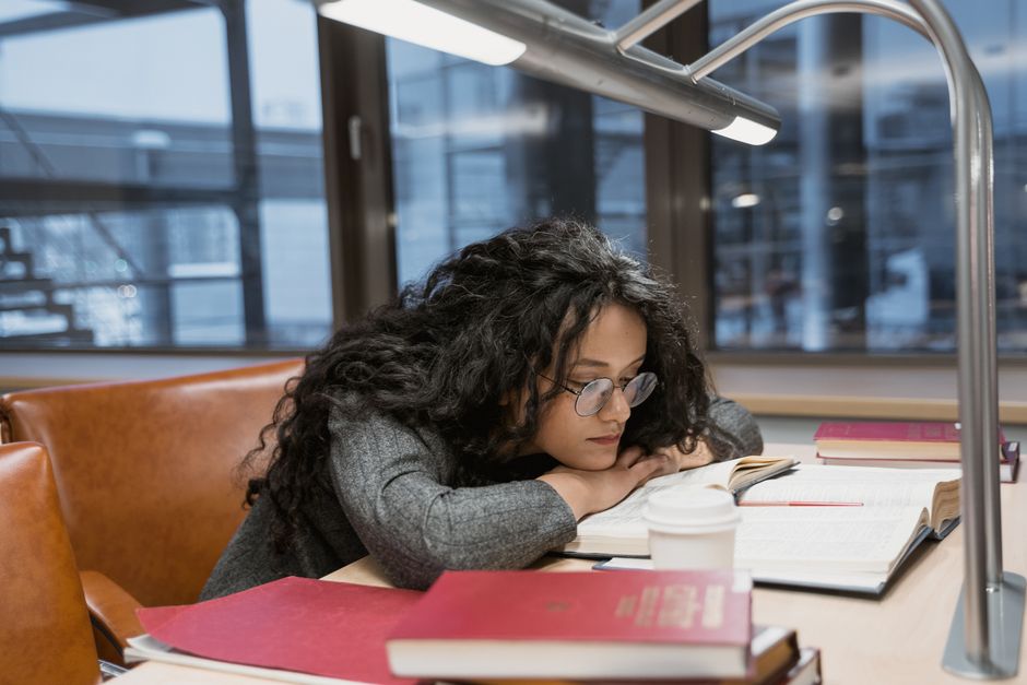 Estudante de estudante cansada, com cabelos cacheados e óculos, descansando a cabeça sobre livros e cadernos em um ambiente de estudo ou biblioteca.