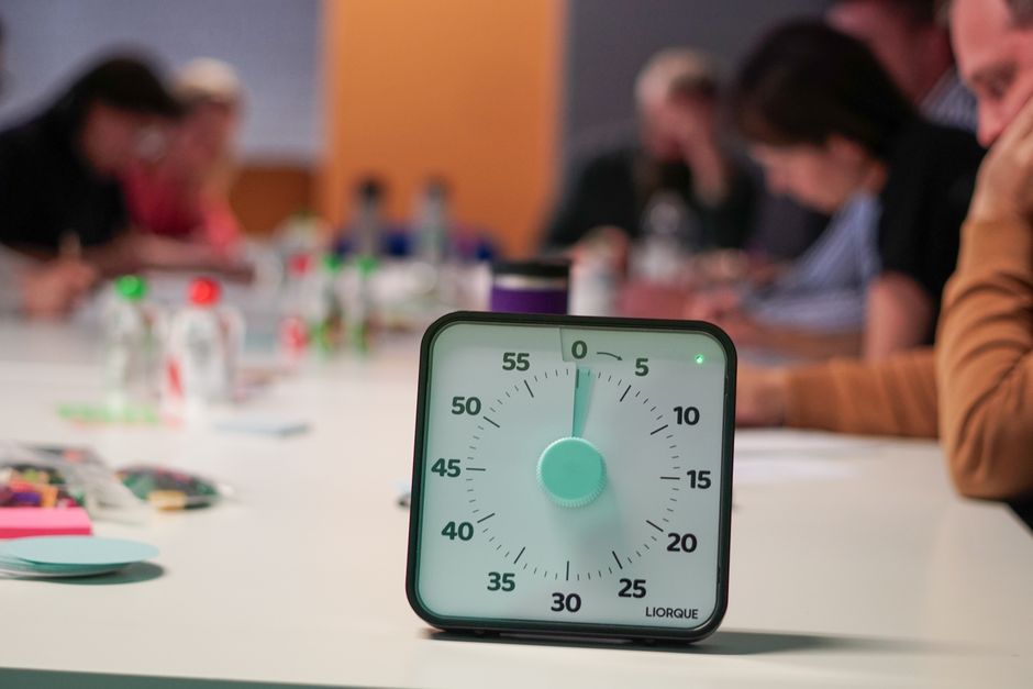 Imagem de um relógio de cozinha com ponteiro na posição zero, simbolizando expectativa de tempo ou início de uma atividade, com pessoas reunidas ao fundo em um ambiente de trabalho ou reunião.