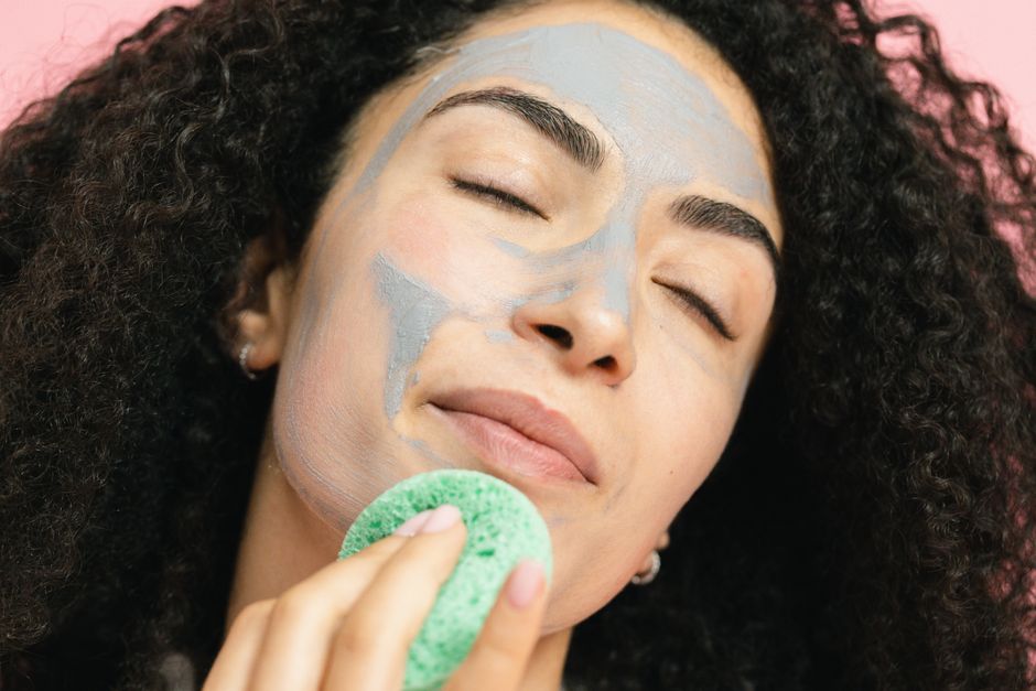 Mulher relaxando e cuidando da pele com uma máscara facial enquanto aplica um removedor com uma esponja verde, evidenciando cuidados de beleza e bem-estar.