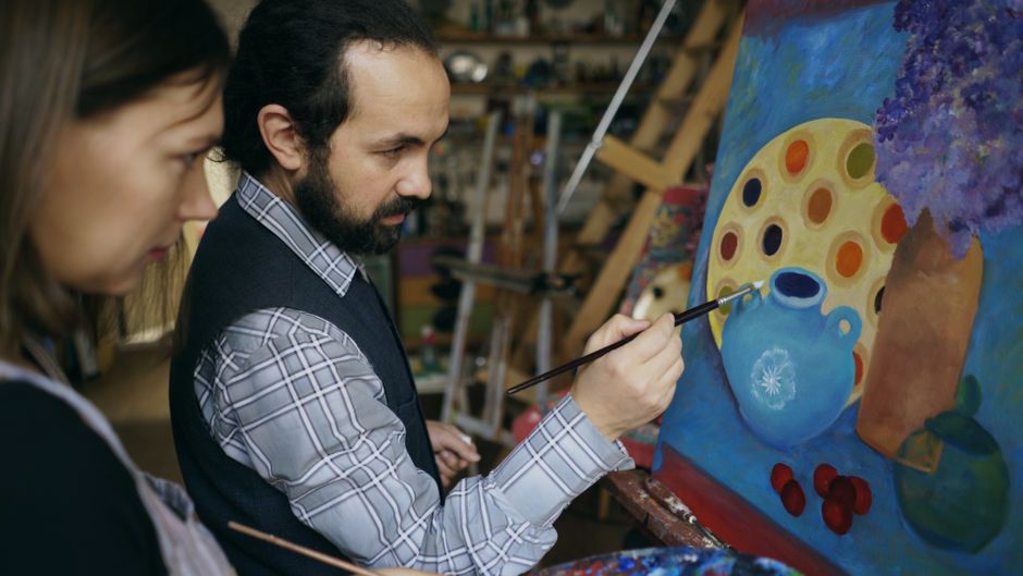 Pintor criando uma obra artística em um ateliê, focado na tela que representa uma composição colorida com utensílios e elementos de natureza morta.