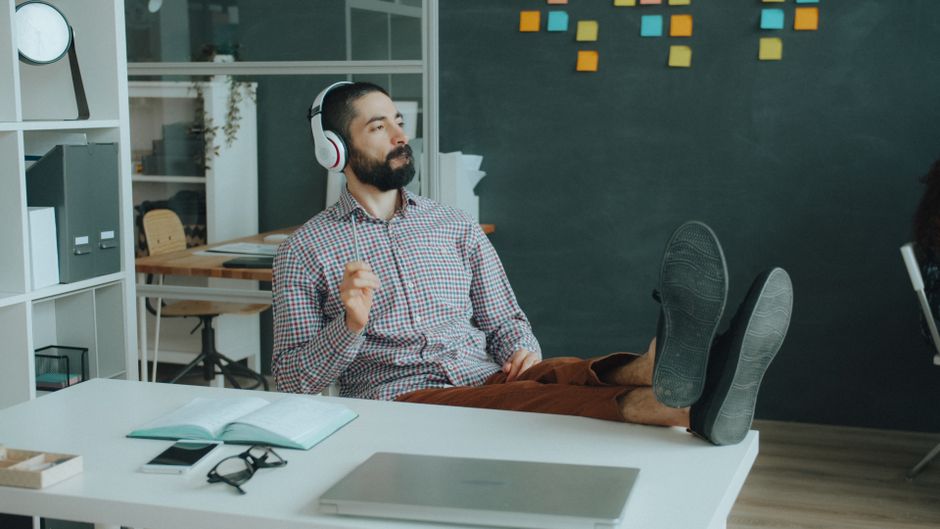 Homem relaxado em ambiente de trabalho moderno, ouvindo música com fones de ouvido e deitado com os pés em cima da mesa, transmitindo sensação de descontração e bem-estar no escritório.