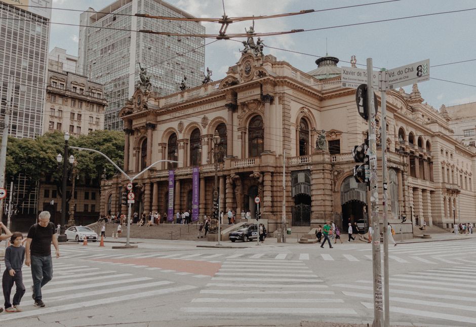 Edifício histórico em um bairro urbano movimentado, com pessoas atravessando a rua, simbolizando a mistura de tradição e modernidade na cidade.