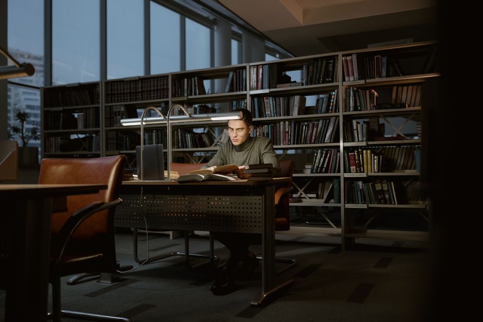 Estudante concentrado estudando em uma biblioteca moderna e bem iluminada com prateleiras cheias de livros ao fundo.