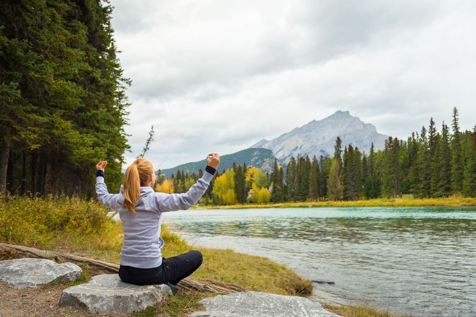 Mulher praticando meditação ao ar livre em meio à natureza com montanhas e rio ao fundo, transmitindo tranquilidade e conexão com o ambiente natural.
