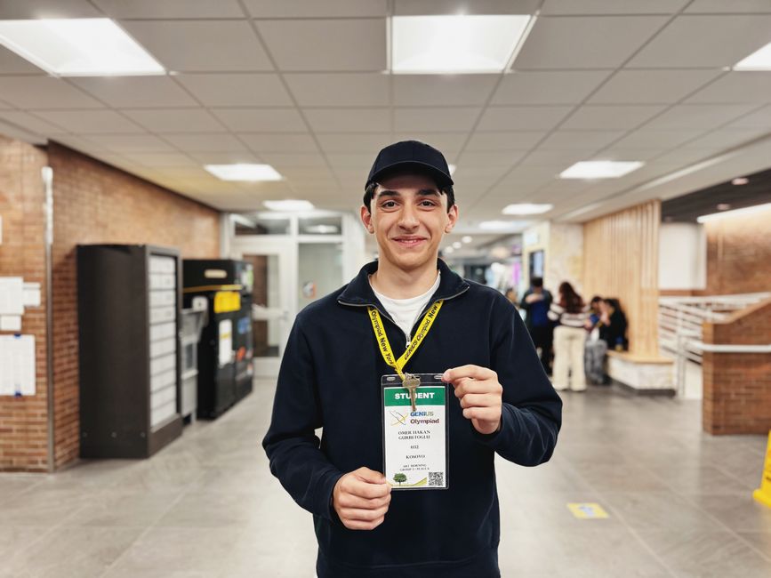 Jovem sorridente exibindo sua credencial de participante em um evento escolar ou competição de ciências, em ambiente interno com colegas ao fundo.
