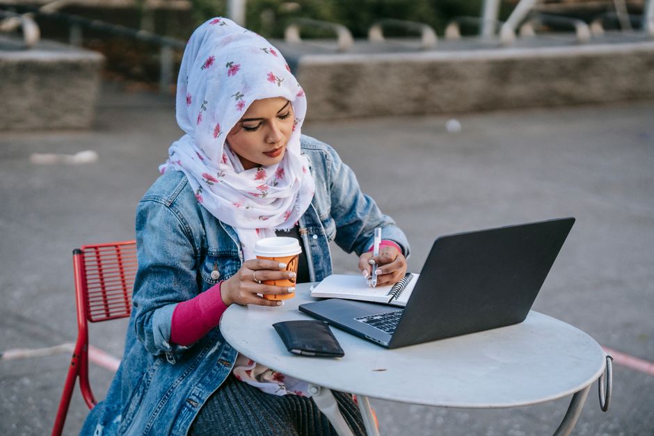 Mulher estudando ou trabalhando ao ar livre, com notebook, caneta, caderno e café em uma mesa, transmitindo produtividade em ambiente externo.