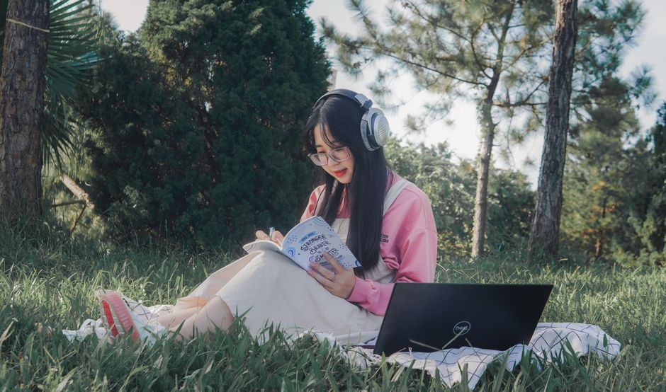 Jovem estudando ao ar livre com fone de ouvido, laptop e livro, em meio à natureza, em um ambiente de estudo ao ar livre.