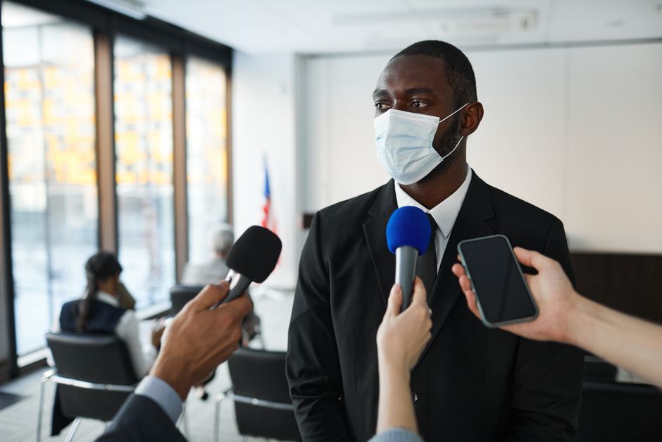 Homem em entrevista de imprensa, usando máscara facial em um ambiente corporativo com janelas amplas e pessoas ao fundo.