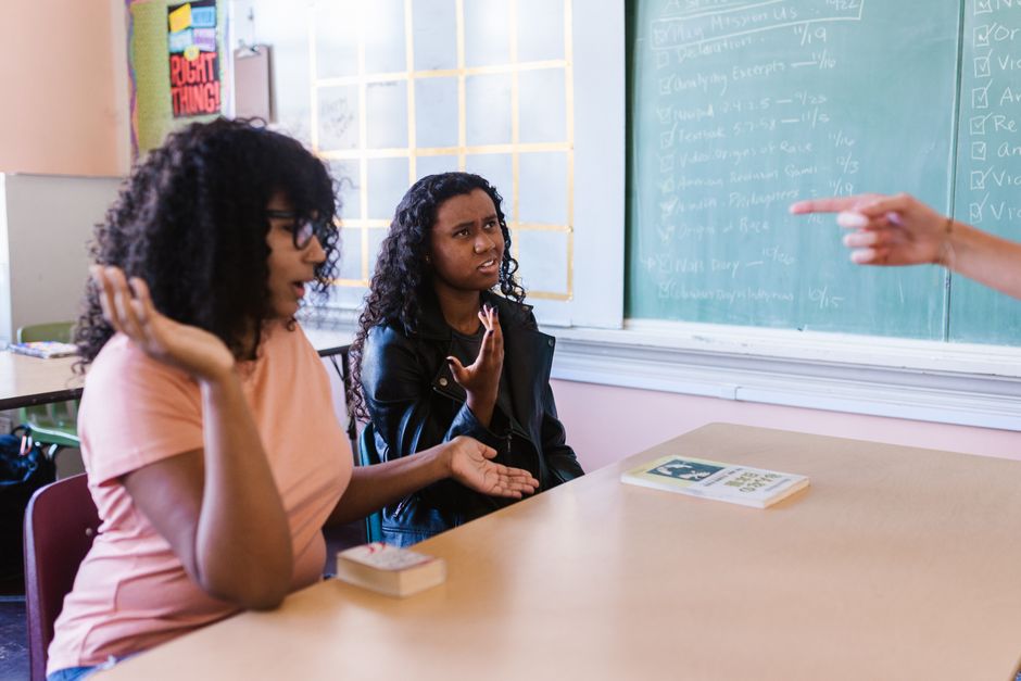 Duas adolescentes discutindo questões escolares em sala de aula, com uma professora apontando um assunto no quadro-negro.