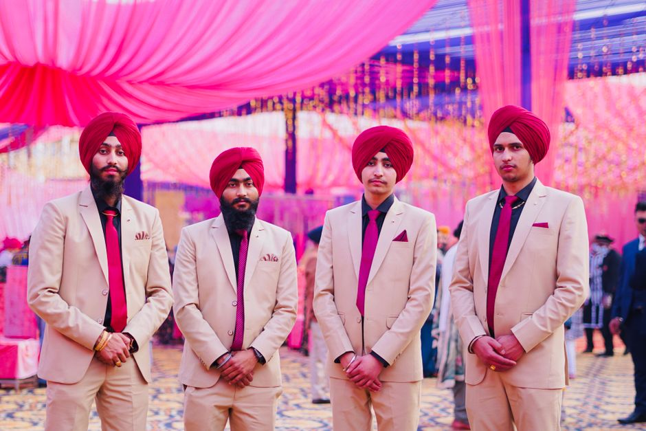 Grupo de homens indianos vestidos com trajes formais e turbantes, participando de um evento cultural ou celebração tradicional sob uma decoração colorida.