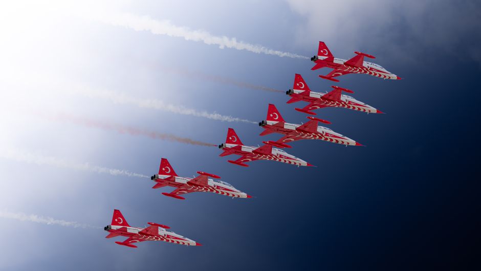 Seis aviões de corrida voando em formação com céu nublado ao fundo, representando velocidade e precisão na aviação esportiva.