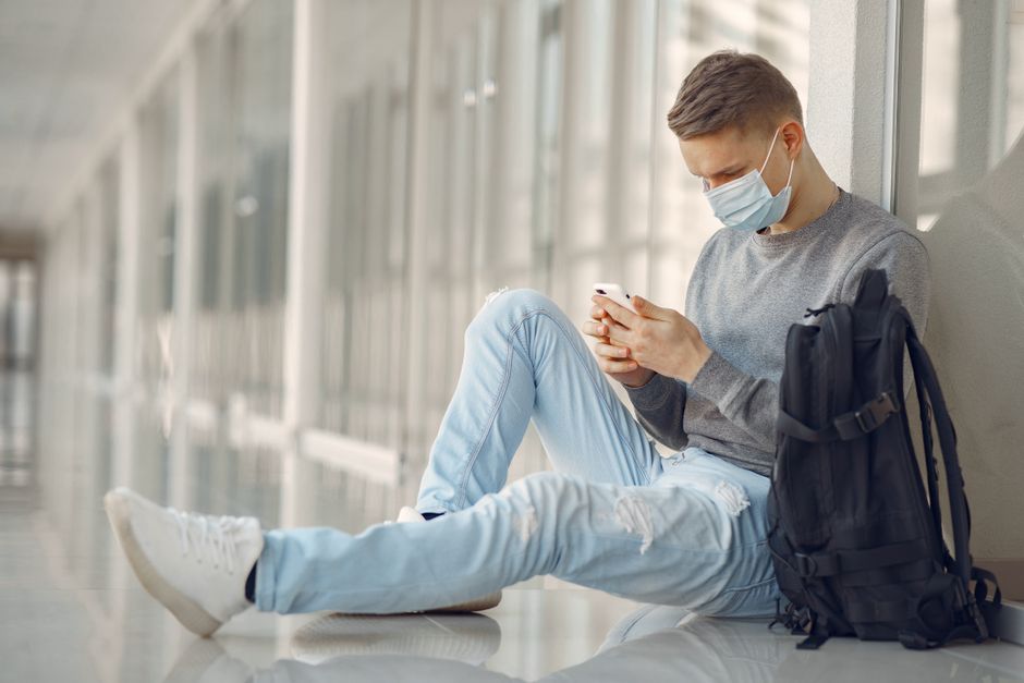 Jovem sentado em ambiente moderno, usando máscara e analisando seu telefone, transportando uma mochila, representando o tema de segurança, saúde e tecnologia na rotina urbana.