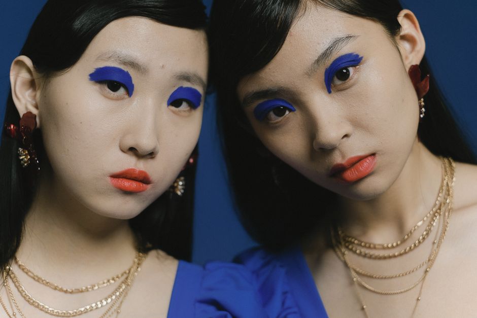 Duas mulheres com maquiagem marcante e joias, em um retrato estilizado com fundo azul, transmitindo moda e expressão artística.